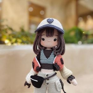 Puede incluir: Una mu&ntilde;eca de crochet que lleva una gorra de b&eacute;isbol blanca y azul con un coraz&oacute;n, un top marr&oacute;n, una chaqueta de crochet beige y pantalones beige con una ri&ntilde;onera negra. La mu&ntilde;eca tiene el pelo marr&oacute;n y una cara de crochet con ojos marrones.