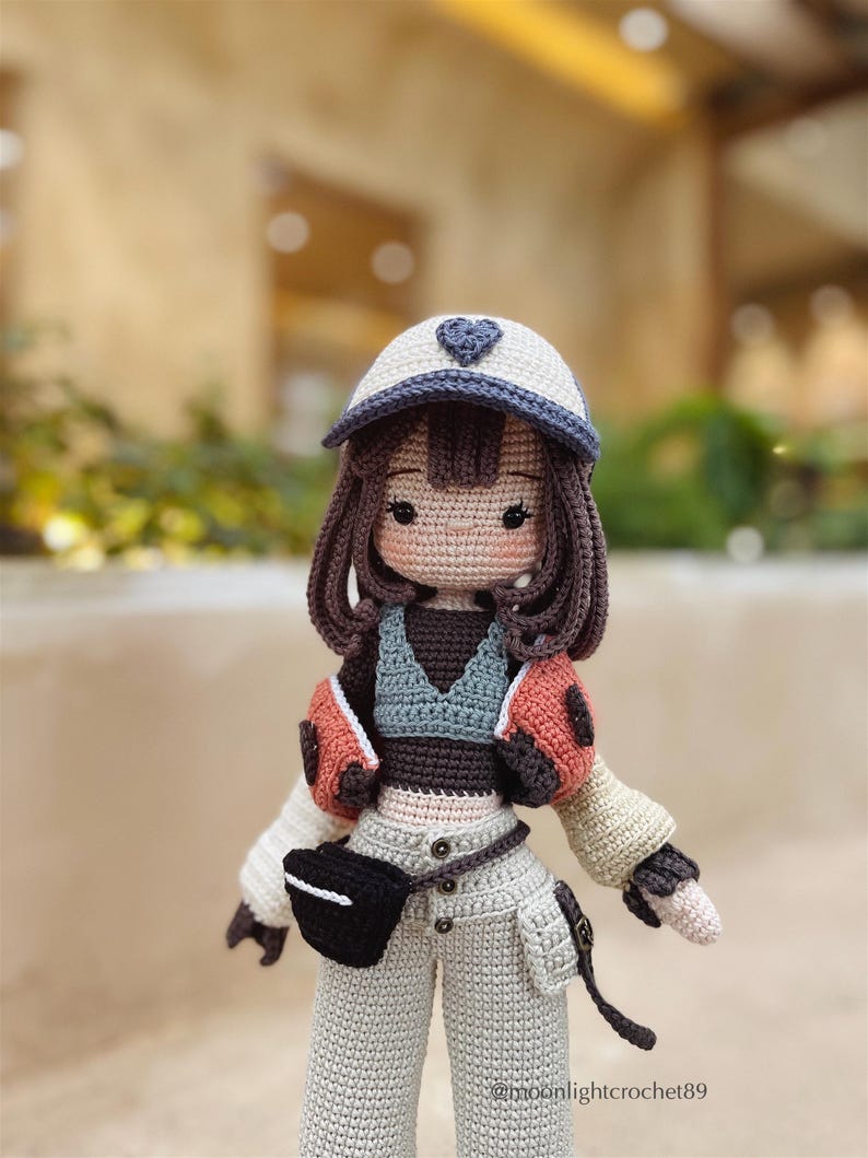 Puede incluir: Una mu&ntilde;eca de crochet que lleva una gorra de b&eacute;isbol blanca y azul con un coraz&oacute;n, un top marr&oacute;n, una chaqueta de crochet beige y pantalones beige con una ri&ntilde;onera negra. La mu&ntilde;eca tiene el pelo marr&oacute;n y una cara de crochet con ojos marrones.