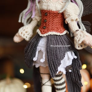 May include: A handmade crochet doll with a detailed design. The doll features a rust-colored bodice, a gray skirt with white accents, and striped stockings. The doll's hair is a mix of gray and purple. The doll is a fairy or a similar fantasy character.