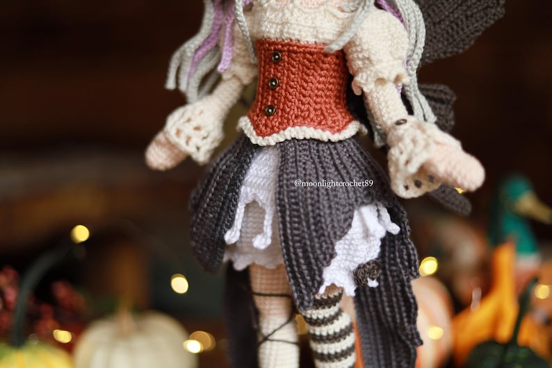 May include: A handmade crochet doll with a detailed design. The doll features a rust-colored bodice, a gray skirt with white accents, and striped stockings. The doll's hair is a mix of gray and purple. The doll is a fairy or a similar fantasy character.