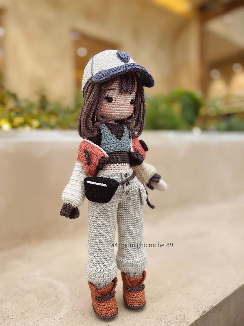 Puede incluir: Una mu&ntilde;eca de crochet con una gorra de b&eacute;isbol blanca, una chaqueta marr&oacute;n y naranja, una camiseta gris, pantalones grises y botas marrones. La mu&ntilde;eca tiene el pelo marr&oacute;n y una cara marr&oacute;n claro. La mu&ntilde;eca est&aacute; de pie sobre una superficie marr&oacute;n. @moonlightcrochet89