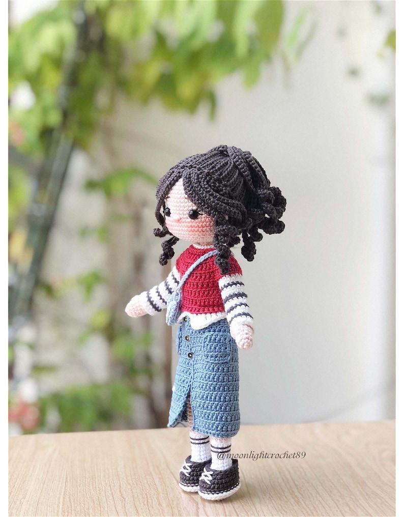 May include: A crocheted doll with brown hair, wearing a red and white striped shirt, a blue denim skirt, and black and white shoes. The doll is standing on a wooden surface. @moonlightcrochet89