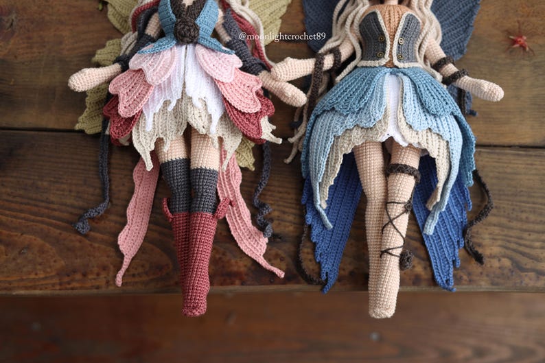 May include: Two handmade crochet fairy dolls with detailed wings and petal-like skirts. One doll features a colour palette of pinks, reds, and greens, while the other showcases blues and tans. Both dolls have long, flowing hair and are posed on a wooden surface.