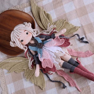 May include: A handmade crochet doll in a fairy design. The doll has a light skin tone, long white hair, and is wearing a dress with pink, white, and blue accents. The doll is lying on a checkered fabric with crochet wings and a wooden plate.