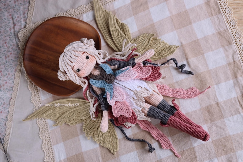May include: A handmade crochet doll in a fairy design. The doll has a light skin tone, long white hair, and is wearing a dress with pink, white, and blue accents. The doll is lying on a checkered fabric with crochet wings and a wooden plate.