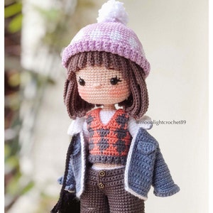 May include: A crocheted doll wearing a purple and white hat, a red and white plaid waistcoat, a blue denim jacket, brown trousers, and a black bag. The doll has brown hair and a white pom-pom on her hat. The doll is wearing a white shirt under the waistcoat. The doll is standing on a white surface. @moolightcrochet89