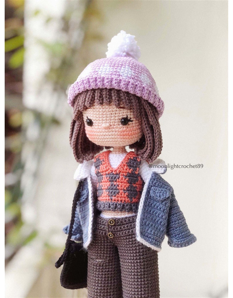 May include: A crocheted doll wearing a purple and white hat, a red and white plaid waistcoat, a blue denim jacket, brown trousers, and a black bag. The doll has brown hair and a white pom-pom on her hat. The doll is wearing a white shirt under the waistcoat. The doll is standing on a white surface. @moolightcrochet89