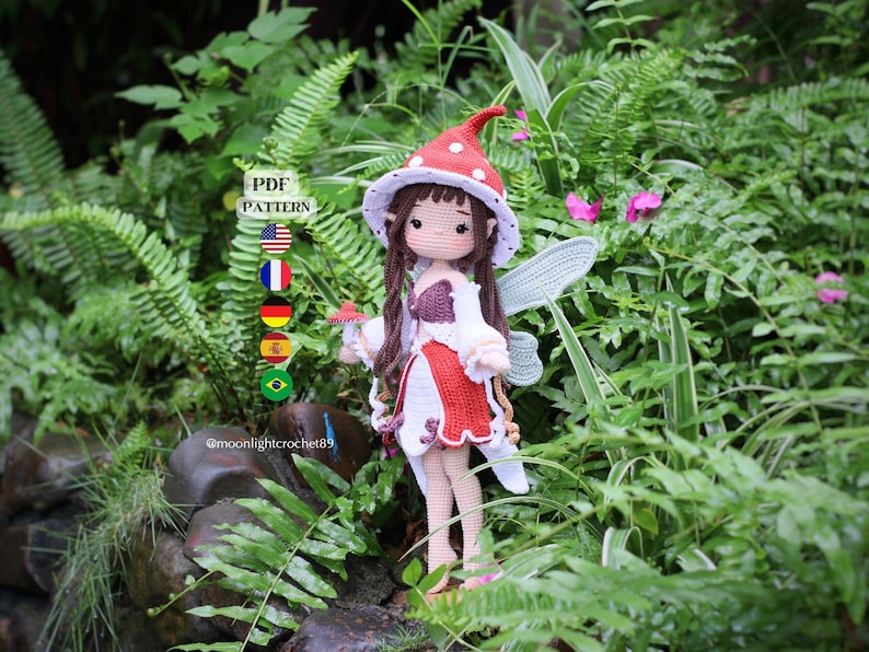 May include: A handmade crochet fairy doll with brown hair, wearing a red and white dress and a mushroom-shaped hat. The doll has wings and is holding a small mushroom. The background features lush green foliage and pink flowers.