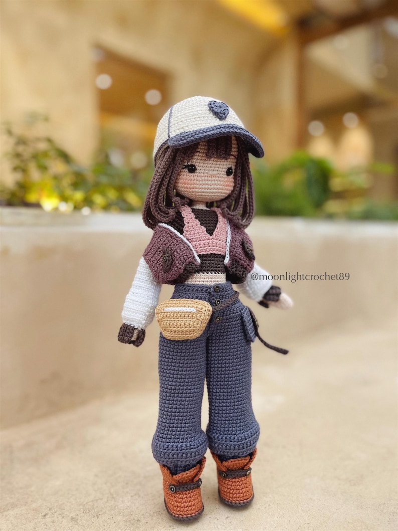 Puede incluir: Mu&ntilde;eca de crochet con una gorra de b&eacute;isbol blanca con un coraz&oacute;n, una chaqueta marr&oacute;n, un top rosa, pantalones vaqueros azules, un cintur&oacute;n marr&oacute;n y botas marrones. La mu&ntilde;eca tiene el pelo marr&oacute;n y una cara de crochet. La mu&ntilde;eca lleva una bolsa de crochet marr&oacute;n. @moonlightcrochet89