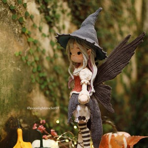 May include: A handcrafted crochet fairy doll with a gray witch hat, wings, and a red and white bodice. The doll is standing on a wooden base, with a skull-shaped lantern. The background includes gourds and fairy lights.