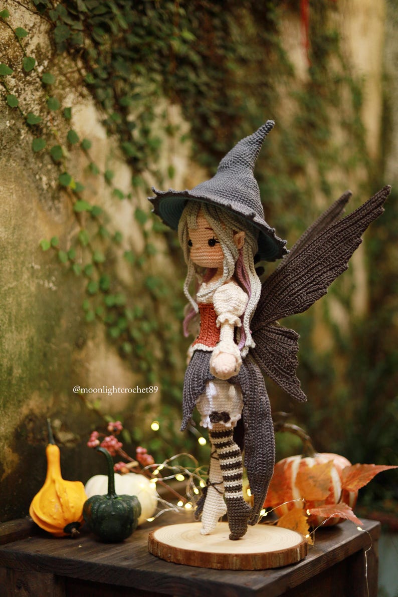 May include: A handcrafted crochet fairy doll with a gray witch hat, wings, and a red and white bodice. The doll is standing on a wooden base, with a skull-shaped lantern. The background includes gourds and fairy lights.