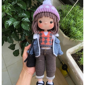 May include: A crocheted doll wearing a purple and white hat, a blue denim jacket, a red and black patterned vest, brown trousers, and black shoes. The doll is holding a black purse.