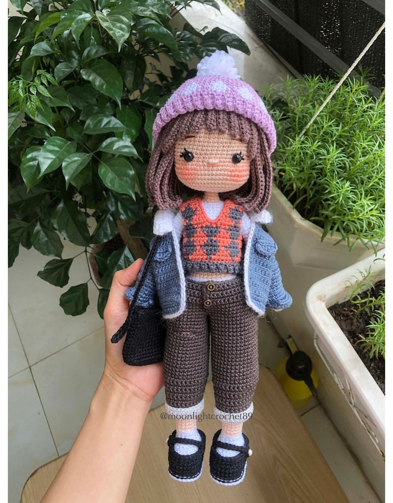 May include: A crocheted doll wearing a purple and white hat, a blue denim jacket, a red and black patterned vest, brown trousers, and black shoes. The doll is holding a black purse.