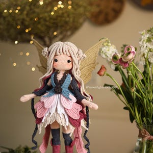 Peut inclure: Une poup&eacute;e au crochet faite &agrave; la main en forme de f&eacute;e. La poup&eacute;e a des cheveux blonds, une robe bleue et rose et des ailes beiges. La poup&eacute;e porte des bas gris fonc&eacute; et des bottes marron. La poup&eacute;e est devant un vase de fleurs.