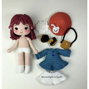 May include: A crocheted doll with brown hair, a white dress, and a blue dress. The doll is surrounded by accessories including a hat, shoes, a purse, and a bib.