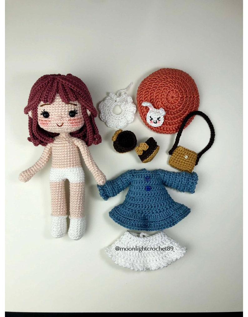 May include: A crocheted doll with brown hair, a white dress, and a blue dress. The doll is surrounded by accessories including a hat, shoes, a purse, and a bib.