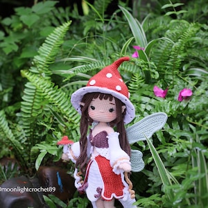 May include: A handmade crochet doll of a fairy with brown hair, wearing a red and white mushroom cap and dress. The doll has blue wings and is holding a small mushroom. The background is lush green foliage.