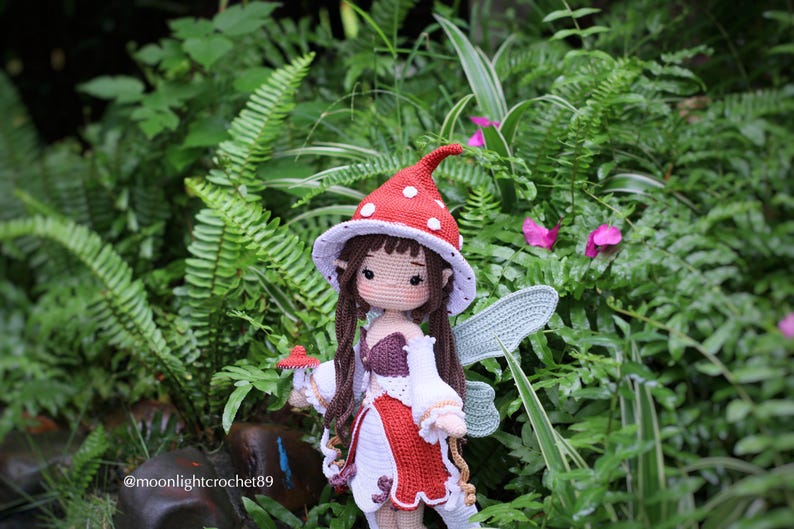 May include: A handmade crochet doll of a fairy with brown hair, wearing a red and white mushroom cap and dress. The doll has blue wings and is holding a small mushroom. The background is lush green foliage.