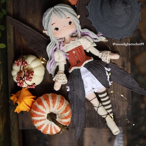 May include: A handmade crochet fairy doll with gray hair and purple highlights, wearing a rust-colored corset and a dark skirt. The doll has brown wings and is surrounded by decorative pumpkins and a witch hat. The text "@moonlightcrochet89" is visible.