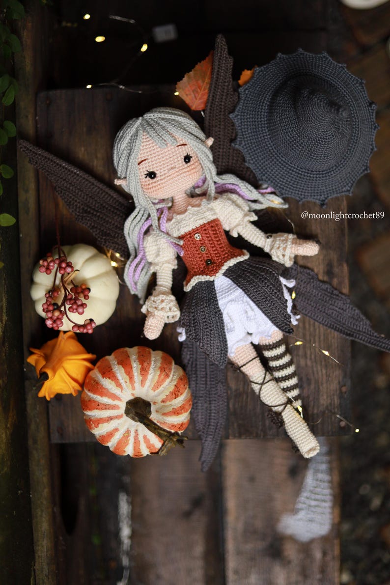 May include: A handmade crochet fairy doll with gray hair and purple highlights, wearing a rust-colored corset and a dark skirt. The doll has brown wings and is surrounded by decorative pumpkins and a witch hat. The text "@moonlightcrochet89" is visible.