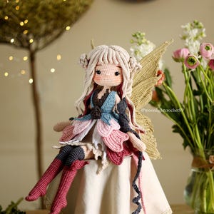 Peut inclure: Une poup&eacute;e de f&eacute;e au crochet faite &agrave; la main avec de longs cheveux blonds, assise sur une surface cr&egrave;me. La poup&eacute;e pr&eacute;sente une robe multicolore avec des accents roses, bleus et blancs, et des ailes dor&eacute;es. Un vase de fleurs roses et blanches est en arri&egrave;re-plan.