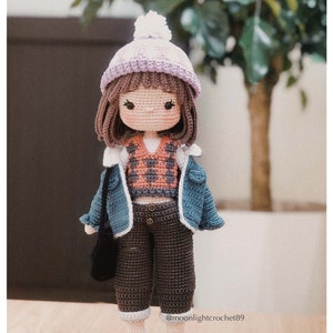 May include: Crocheted doll wearing a purple and white hat, a blue denim jacket, a brown vest, brown trousers, and black shoes. The doll is holding a black bag.