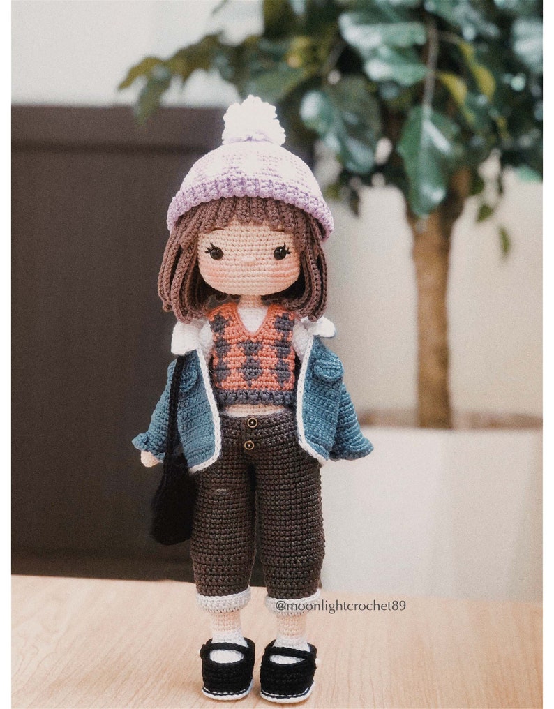 May include: Crocheted doll wearing a purple and white hat, a blue denim jacket, a brown vest, brown trousers, and black shoes. The doll is holding a black bag.
