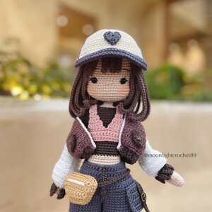 Puede incluir: Mu&ntilde;eca de crochet con cabello casta&ntilde;o que lleva una gorra de b&eacute;isbol blanca y azul con un coraz&oacute;n, un top de crochet rosa y marr&oacute;n, una chaqueta de crochet marr&oacute;n, un cintur&oacute;n marr&oacute;n con una bolsa de crochet marr&oacute;n y pantalones de crochet azules. La mu&ntilde;eca sostiene una bolsa de crochet marr&oacute;n.