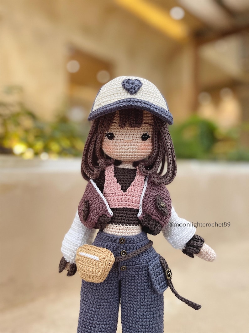Puede incluir: Mu&ntilde;eca de crochet con cabello casta&ntilde;o que lleva una gorra de b&eacute;isbol blanca y azul con un coraz&oacute;n, un top de crochet rosa y marr&oacute;n, una chaqueta de crochet marr&oacute;n, un cintur&oacute;n marr&oacute;n con una bolsa de crochet marr&oacute;n y pantalones de crochet azules. La mu&ntilde;eca sostiene una bolsa de crochet marr&oacute;n.