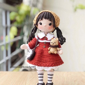 May include: A crocheted doll wearing a red dress, a white collar, a brown hat, and brown shoes. The doll is holding a small crocheted teddy bear. The doll has brown hair and brown eyes. The doll is standing on a wooden surface. @moonlightcrochet89