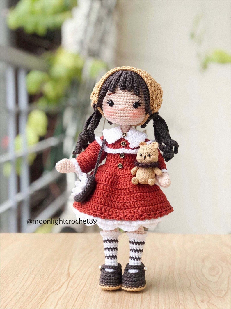 May include: A crocheted doll wearing a red dress, a white collar, a brown hat, and brown shoes. The doll is holding a small crocheted teddy bear. The doll has brown hair and brown eyes. The doll is standing on a wooden surface. @moonlightcrochet89