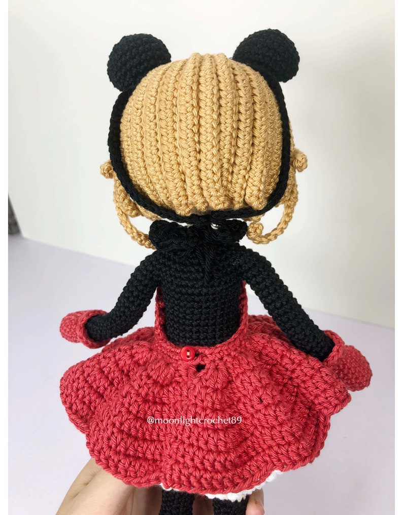 K&ouml;nnte beinhalten: Geh&auml;kelte Puppe mit schwarzen B&auml;renohren, einem schwarzen Kleid und einem roten Rock. Die Puppe tr&auml;gt einen roten Knopf auf der R&uuml;ckseite des Kleides. Die Puppe h&auml;lt ihre Arme seitlich ausgestreckt. Die Puppe steht auf einer wei&szlig;en Oberfl&auml;che. Der Text "@moonlightcrochet89" ist auf dem Rock sichtbar.