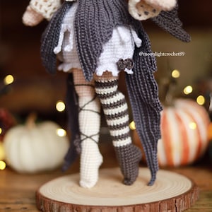 May include: Crocheted doll with a dark gray and white outfit, including a skirt and striped stockings. The doll is standing on a wooden slice, with pumpkins and decorative lights in the background. The doll's outfit has a witchy theme.
