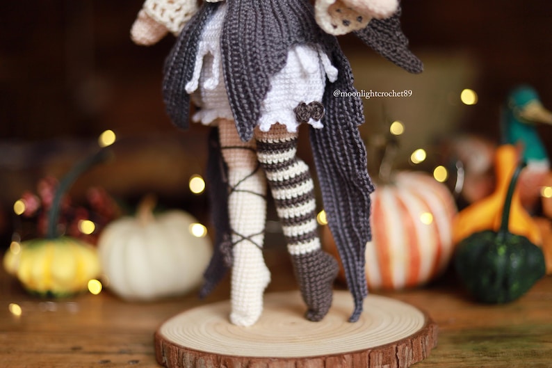 May include: Crocheted doll with a dark gray and white outfit, including a skirt and striped stockings. The doll is standing on a wooden slice, with pumpkins and decorative lights in the background. The doll's outfit has a witchy theme.