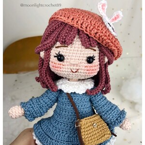 May include: Crocheted doll with brown hair, wearing a blue dress, a brown satchel, and a pink beret with a white bunny ear accent.  The doll has a sweet smile and large black eyes.