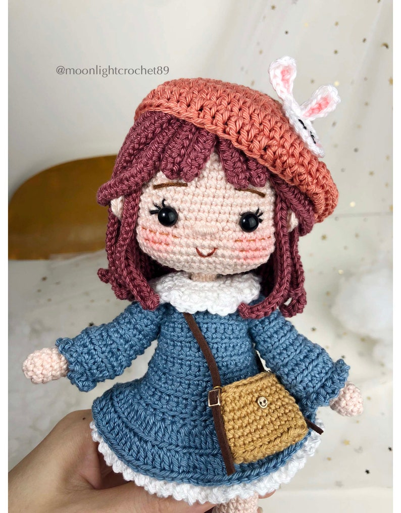 May include: Crocheted doll with brown hair, wearing a blue dress, a brown satchel, and a pink beret with a white bunny ear accent.  The doll has a sweet smile and large black eyes.