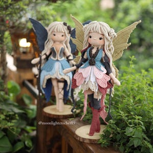 May include: Two handmade crochet fairy dolls with blonde hair and detailed wings. One doll wears a blue and black dress, the other a pink, blue, and white dress. Both dolls stand on wooden bases, surrounded by greenery.