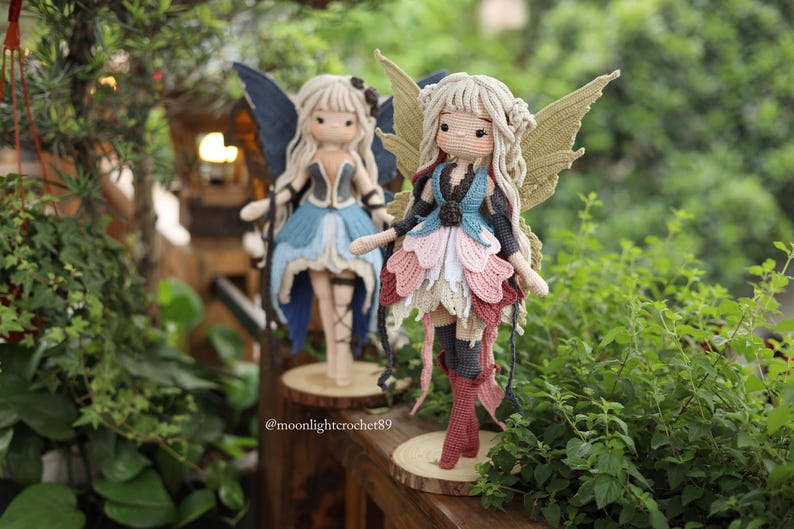 May include: Two handmade crochet fairy dolls with blonde hair and detailed wings. One doll wears a blue and black dress, the other a pink, blue, and white dress. Both dolls stand on wooden bases, surrounded by greenery.
