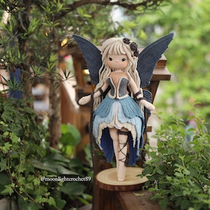 May include: A handcrafted crochet fairy doll with long blonde hair, blue wings, and a blue and beige dress. The doll is standing on a wooden slice, surrounded by greenery. The doll's wings are a dark blue colour.