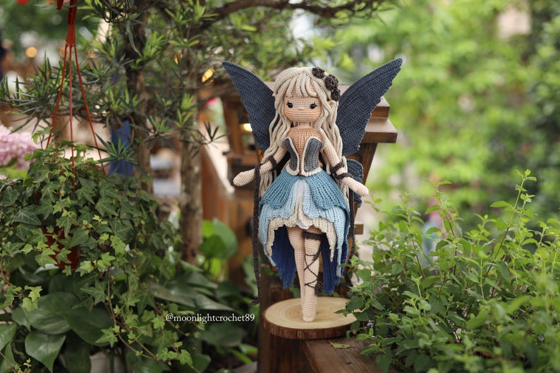 May include: A handcrafted crochet fairy doll with long blonde hair, blue wings, and a blue and beige dress. The doll is standing on a wooden slice, surrounded by greenery. The doll's wings are a dark blue colour.