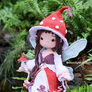 May include: A handmade crochet fairy doll with a red mushroom cap hat with white spots. The doll has brown hair, a white and red dress, and blue wings. The doll is holding a small mushroom.