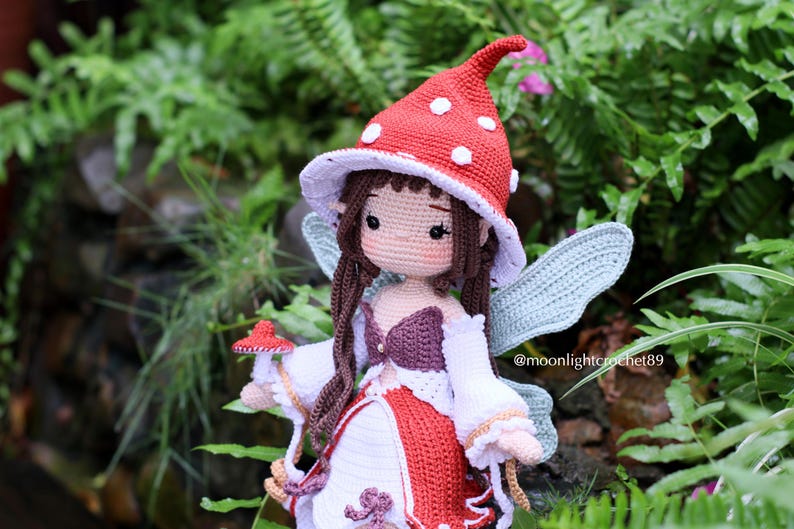 May include: A handmade crochet fairy doll with a red mushroom cap hat with white spots. The doll has brown hair, a white and red dress, and blue wings. The doll is holding a small mushroom.
