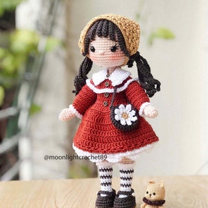 May include: A crocheted doll wearing a red dress, white collar, brown shoes, and a brown and beige hat. The doll has brown hair in pigtails and is holding a white crocheted purse with a flower design. A small crocheted brown teddy bear is sitting in front of the doll.