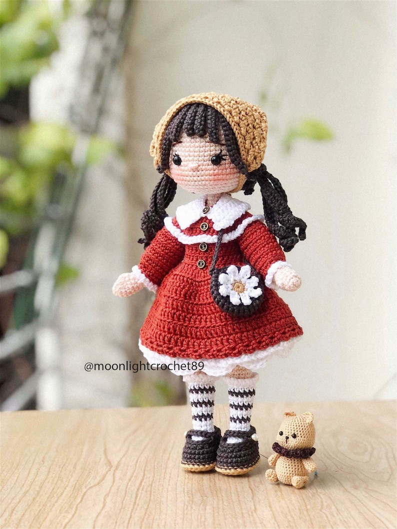May include: A crocheted doll wearing a red dress, white collar, brown shoes, and a brown and beige hat. The doll has brown hair in pigtails and is holding a white crocheted purse with a flower design. A small crocheted brown teddy bear is sitting in front of the doll.