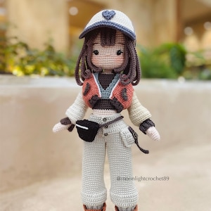 Puede incluir: Una mu&ntilde;eca de crochet que lleva un sombrero blanco y azul con un coraz&oacute;n, un su&eacute;ter marr&oacute;n y naranja, pantalones blancos, una bolsa negra y botas marrones. La mu&ntilde;eca tiene el pelo marr&oacute;n y una cara de crochet con ojos marrones.