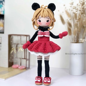 K&ouml;nnte beinhalten: Eine geh&auml;kelte Puppe mit schwarzer und roter Kleidung, einer wei&szlig;en Schleife und schwarzen M&auml;useohren. Die Puppe hat blonde Haare und tr&auml;gt rote Schuhe.
