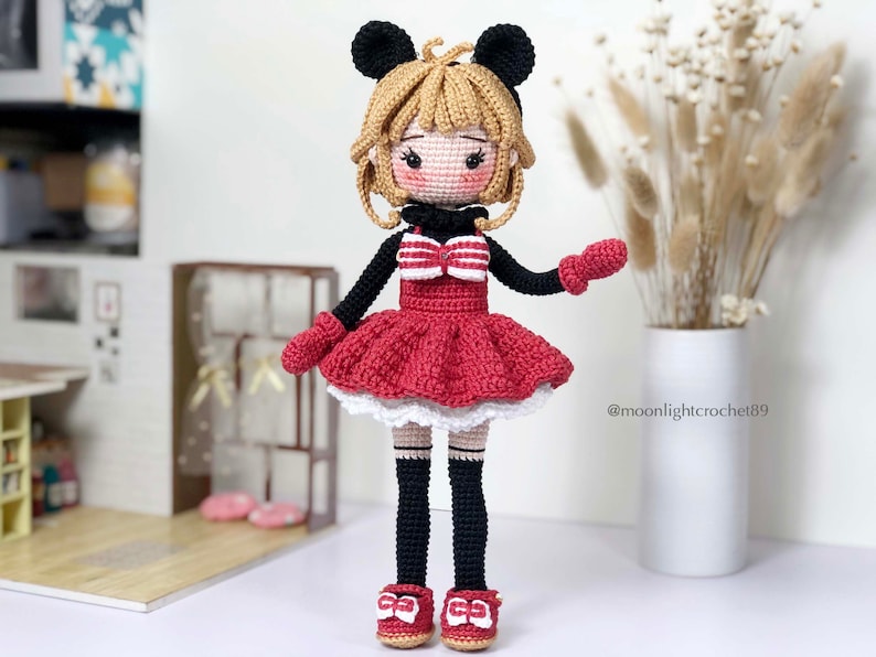 K&ouml;nnte beinhalten: Eine geh&auml;kelte Puppe mit schwarzer und roter Kleidung, einer wei&szlig;en Schleife und schwarzen M&auml;useohren. Die Puppe hat blonde Haare und tr&auml;gt rote Schuhe.