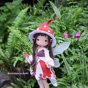 May include: A handmade crochet fairy doll with a red and white mushroom cap hat, wings, and dress. The doll has long brown hair and is holding a small mushroom. The background is lush green foliage.
