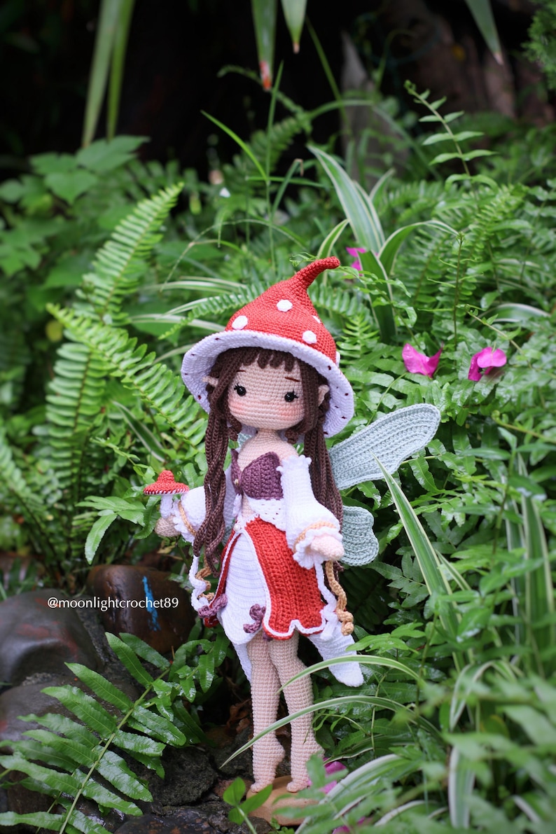 May include: A handmade crochet fairy doll with a red and white mushroom cap hat, wings, and dress. The doll has long brown hair and is holding a small mushroom. The background is lush green foliage.