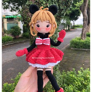 K&ouml;nnte beinhalten: Eine geh&auml;kelte Puppe mit schwarzen M&auml;useohren, einem roten Kleid mit wei&szlig;em Besatz, schwarzen Leggings und roten Schuhen. Die Puppe hat blondes Haar und eine rote Schleife. Die Puppe h&auml;lt einen roten F&auml;ustling in ihrer rechten Hand. Die Puppe sitzt auf einer Hand. Die Puppe wurde von @moonlightcrochet89 hergestellt.
